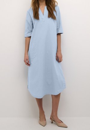 Femme portant une robe bleu clair mi-longue avec manches trois-quarts et chaussures beige à talons bas pointues, debout devant un fond uni.