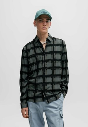 Black and gray long-sleeve shirt with a grid pattern, made of a smooth fabric. Model wears a light blue cap and cargo pants.