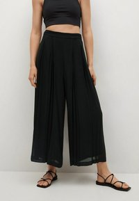 Zwarte wijde broek met plooien, hoge taille, vloeiende stof en transparante textuur. Model draagt zwarte sandaaltjes met bandjes.