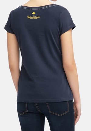 Kobieta ubrana w granatowy t-shirt z krótkim rękawem z żółtym logo i napisem na górnej części pleców, w połączeniu z ciemnoniebieskimi jeansami.