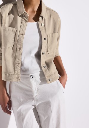 Personne portant une veste beige à boutons par-dessus une chemise blanche et un pantalon blanc, avec une main dans la poche et un fond neutre.