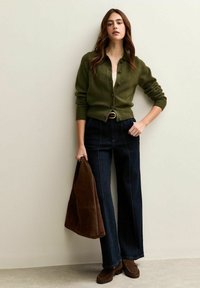 Mujer con cárdigan verde oliva abotonado, jeans oscuros de cintura alta, mocasines marrones, lleva un bolso de ante marrón, de pie contra una pared clara.