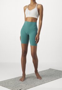 Conjunto atlético que incluye un sujetador deportivo gris claro y unos pantalones cortos de ciclismo de tiro alto en verde azulado, en una modelo que está descalza sobre una esterilla gris texturizada.
