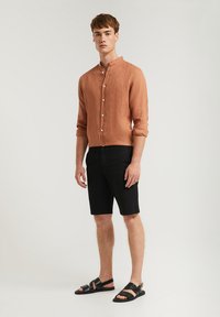 Bränd orange linneskjorta med uppvikta ärmar och knäppning, tillsammans med svarta shorts och svarta sandaler, på en neutral bakgrund.