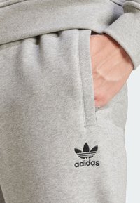 Šedé tepláky vyrobené z měkké látky, s vyšitým černým logem Adidas a kapsami s akcentem. Volný střih s elastickým pasem.