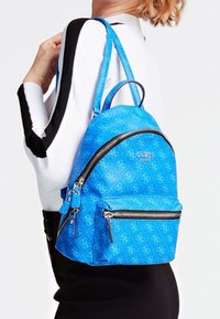 Niebieski syntetyczny plecak o zaokrąglonym kształcie, z wzorem logo 'Guess', srebrnymi elementami metalowymi oraz przednią kieszenią na zamek.