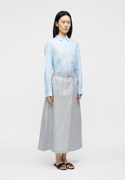 Γυναίκα στέκεται φορώντας ένα ανοιχτό μπλε πουκάμισο με κουμπιά, ριγέ φούστα μέχρι τον αστράγαλο με τσέπες και μαύρα σανδάλια τύπου slip-on σε απλό φόντο.