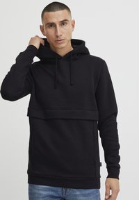 Sudadera con capucha negra hecha de tejido suave, que cuenta con un bolsillo frontal, un diseño en capas y cordones ajustables en la capucha.