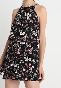 Kvinna i en ärmlös svart blommig sommarklänning med en samlad halterneck och ett knytliv.