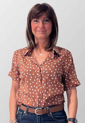 Camicia marrone a pois con maniche corte, chiusura frontale a bottoni e colletto. Indossata con una cintura marrone e pantaloni in denim. Texture liscia.
