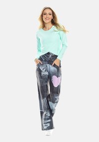 Hellgrünes langärmliges Oberteil kombiniert mit schwarzen Jeanshosen, die mit einem rosa Herz-Patch und Distressed-Details versehen sind. Weiße Sneakers runden den Look ab.