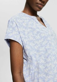 Blusa azzurro chiaro con maniche corte, motivo floreale bianco, design a V, tessuto morbido, dotata di una piccola tasca sul petto.