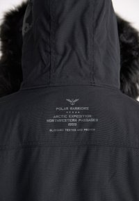 Svart hooded jacka med fuskpälsdetalj, med präglad text som detalj på märket och expeditionens historia på baksidan. Texturerad tyg.