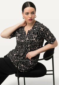 Top negro con estampado abstracto de letras beige, de mangas cortas y con escote en V. Modelo sentado en una silla negra, mostrando una pose relajada.