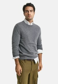 Un pull en laine gris à col rond superposé sur une chemise blanche à col, associé à un pantalon cargo olive avec des poches et une ceinture marron.