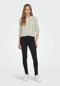 Helles beige Strickoberteil mit offenem Muster, cropped Design und strukturierten Akzenten, kombiniert mit schwarzen Skinny-Jeans und weißen Sneakers.