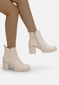 Botas de tobillo beige con tacón grueso, textura suave, cremallera lateral y una pestaña para facilitar el calzado. Suela con relieve para mejorar el agarre.