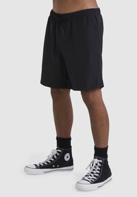 Shorts de bain noirs avec une taille élastique, cordon de serrage et patch logo. Portés avec des baskets montantes noires et des chaussettes mi-mollet.