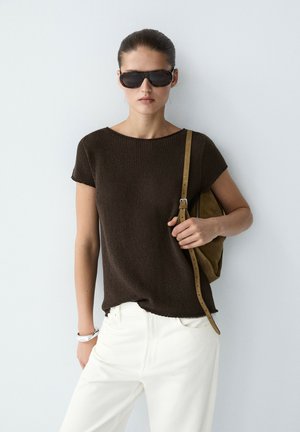 Giovane donna che indossa occhiali da sole neri, top a maniche corte marrone scuro, pantaloni bianchi e che tiene una tracolla marrone di una borsa su uno sfondo semplice.