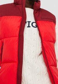 Hombre vistiendo una chaqueta de pluma roja desabotonada sobre un suéter de punto blanco con un texto negro parcialmente visible en el pecho.