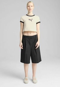 Beige cropped t-shirt med sort ribbet krave, med et flerfarvet logo, parret med sorte plisserede shorts og lysegrå slip-on sko.