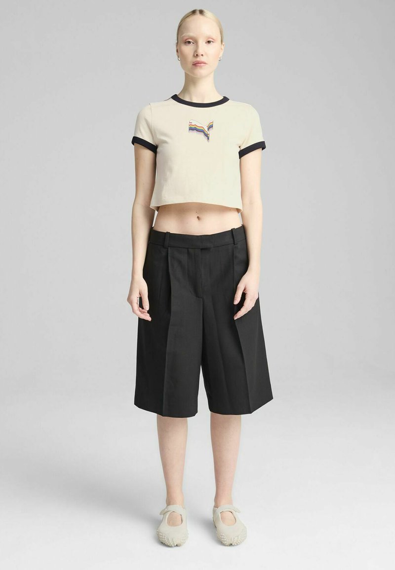 Beige cropped t-shirt med sort ribbet krave, med et flerfarvet logo, parret med sorte plisserede shorts og lysegrå slip-on sko.