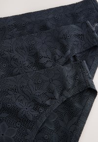 Tissu en dentelle noire à motifs floraux, montrant une texture tissée détaillée et des bords qui se chevauchent sur un fond clair.