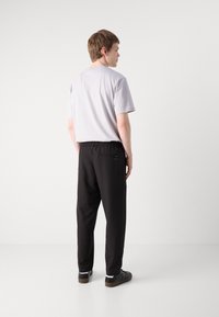T-shirt gris à manches courtes et pantalon noir à taille élastique. Le pantalon a une poche arrière avec un détail de logo. Le modèle se tient de dos, tourné vers le spectateur.