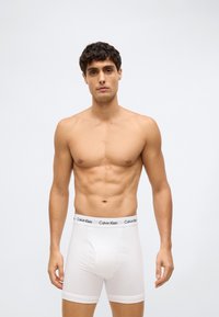 Muž s krátkými tmavými vlasy, oblečený v bílých boxerkách Calvin Klein, stojící proti jednoduchému světlému šedému pozadí.
