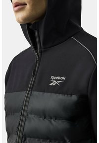 Czarna sportowa kurtka z zamkiem z przodu, kapturem i puchowym dolnym odcinkiem. Posiada srebrne logo Reebok oraz kontrastujące tekstury.