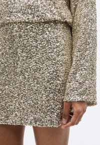 Μικρό χρυσό φόρεμα με παγιέτες και μακριά μανίκια, με υφή από λαμπερές παγιέτες που αντανακλούν το φως με πολυχρωμία.