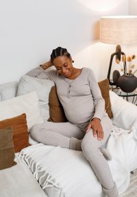 Lichtgrijze, geribbelde loungewearset met lange mouwen en broek, gecombineerd met zachte bruine en crème kussens op een textuurrijke bank. Warme verlichting.