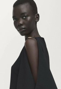 Donkerharig persoon met kort haar, gekleed in een mouwloos zwart kledingstuk, kijkt iets opzij tegen een effen lichte achtergrond.