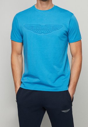 T-shirt di cotone blu con scollo rotondo, caratterizzata da un logo Aston Martin in rilievo sul petto. Abbinata a pantaloni blu navy.