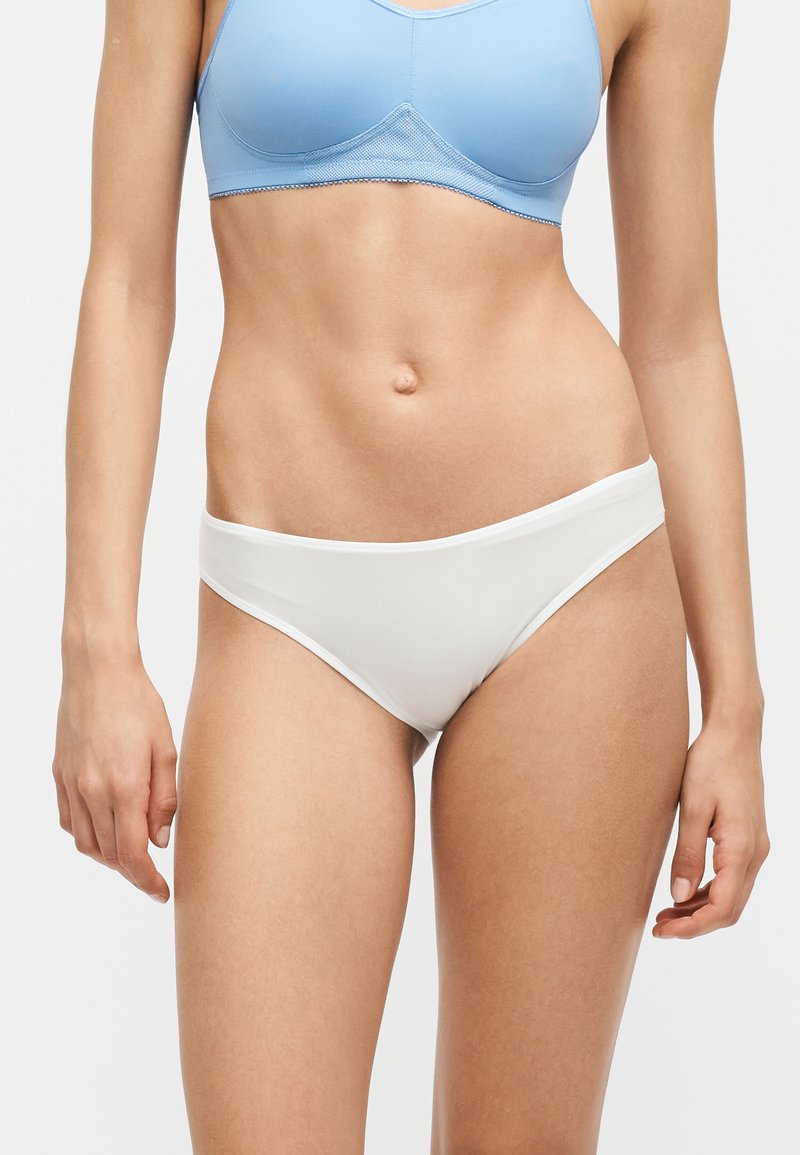 Femme portant un soutien-gorge bleu clair et un sous-vêtement blanc, montrée du milieu du torse jusqu'aux cuisses supérieures sur un fond uni.