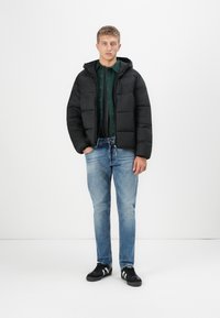 Sort pufferjakke med hætte, quilteret design. Mørkegrøn skjorte med knapper lagdelt under, blå jeans og sorte sneakers med hvide striber.