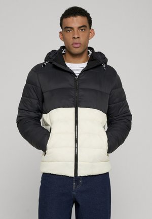 Mann trägt eine schwarz-weiße Daunenjacke mit Kapuze, steht mit den Händen in den Taschen vor einem schlichten grauen Hintergrund.