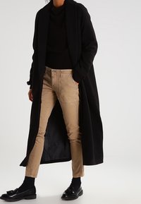 Manteau long noir avec une texture lisse, porté sur un pull noir et un pantalon beige. La tenue comprend des chaussures noires et des chaussettes sombres.