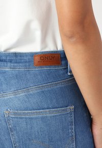Denimjeans i lys blå med et lærremsemerke merket "ONLY." Funksjoner inkluderer baklommer og synlige sømdetaljer.