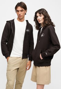 Jeune homme et femme portant des sweats à capuche noirs zippés et des pantalons beiges, debout côte à côte devant un fond uni.