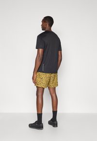 Camiseta preta de mangas curtas, combinada com calções amarelos com padrão de leopardo. Meias e sapatos pretos completam o conjunto. Design simples e funcional.