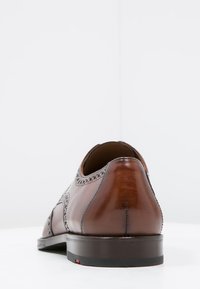 Scarpa da uomo in pelle marrone con dettagli brogue, texture liscia, punta arrotondata e tacco basso nero. Design minimalista con accenti classici.
