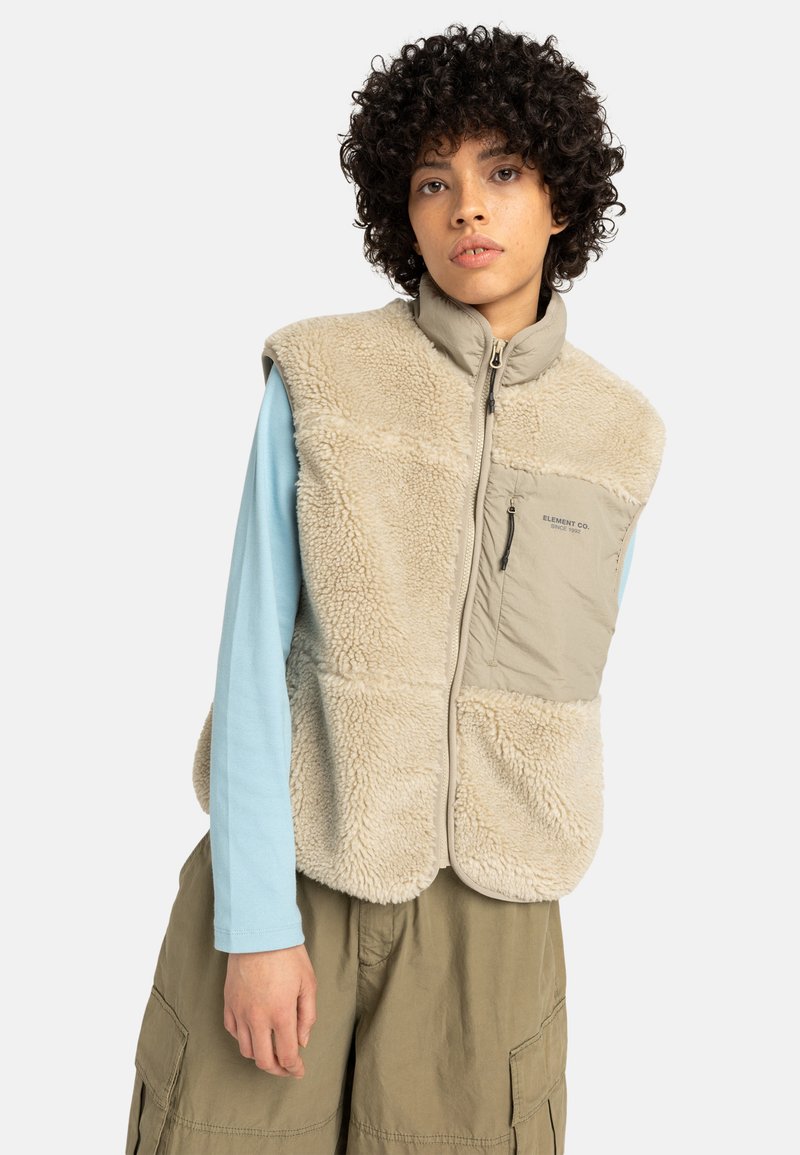 En ung person med lockigt hår bär en beige fleeceväst över en ljusblå långärmad skjorta, stående mot en vit bakgrund.
