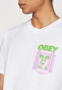 Baltas medvilninis marškinėliai su priekiniais grafikos elementais, kuriuose yra žaliai ir rožinėmis spalvomis užrašas "OBEY" bei tvora, su fraze "BREAKING CHAINS WORLDWIDE."