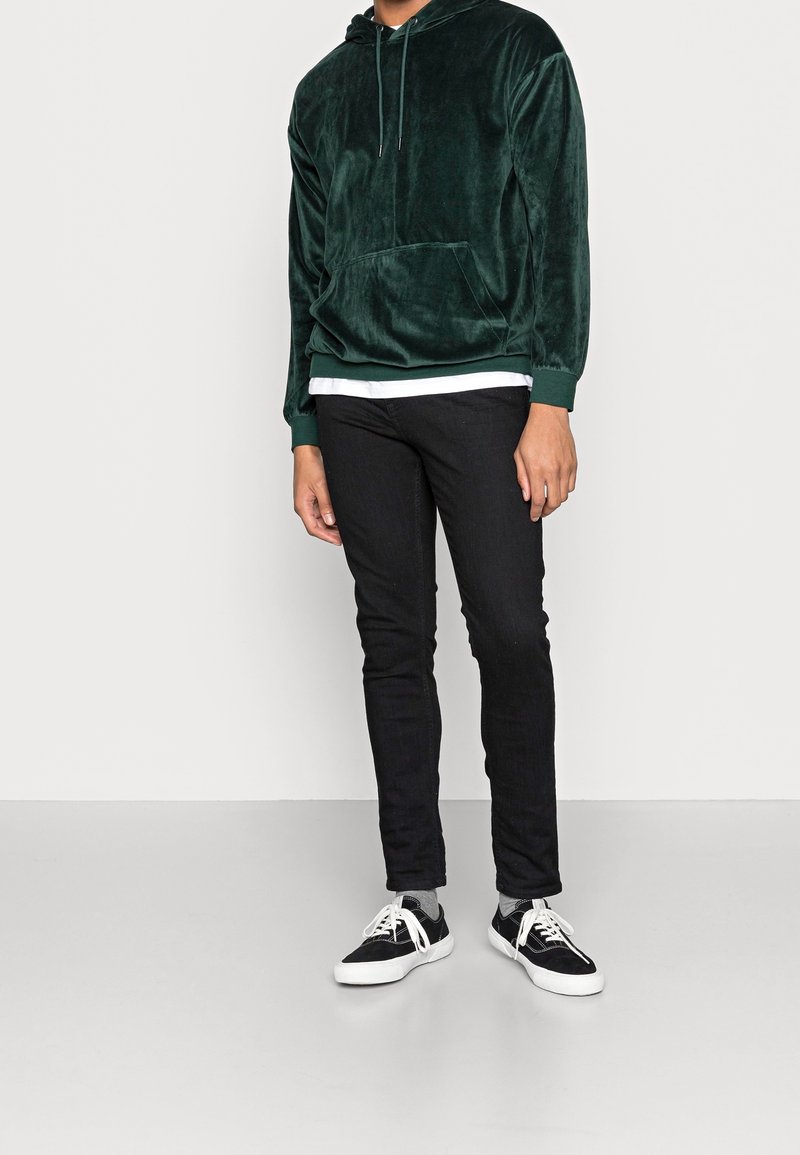 Grön sammetsliknande hoodie med en ficka framtill och dragsko i huvan, kombinerad med svarta slim-fit jeans och svartvita sneakers.