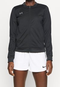 Black zip-up athletic jacket made of smooth fabric with long sleeves, ribbed cuffs, and a subtle logo on the chest. Paired with white shorts.