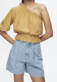 Top mostaza amarillo con un solo hombro y escote con volantes, combinado con shorts de mezclilla azul claro de cintura alta, con pliegues y corte holgado.