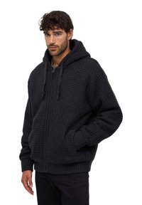 Czarna dzianinowa bluza z kapturem na zamek błyskawiczny o teksturowanym wykończeniu, przestronny kaptur oraz ściągacze na rękawach i dole. Model nosi ją w luźnym fasonie.