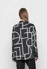Camisa negra con grandes patrones geométricos en blanco, mangas largas, cuello con solapa y un corte relajado. Tejido suave con un ligero brillo.