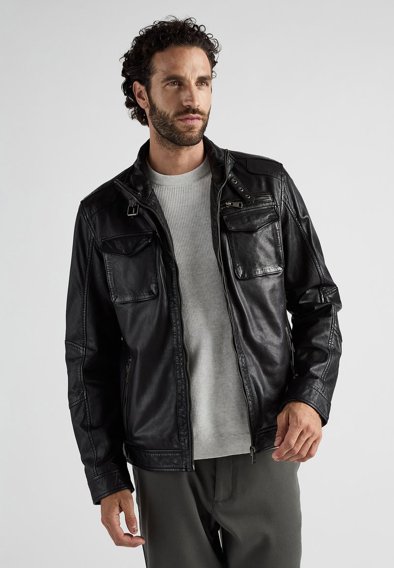 Schwarze Lederjacke mit Reißverschluss, zwei Brusttaschen und Schulterdetails. Getragen über einem hellgrauen Pullover und olivgrünen Hosen.